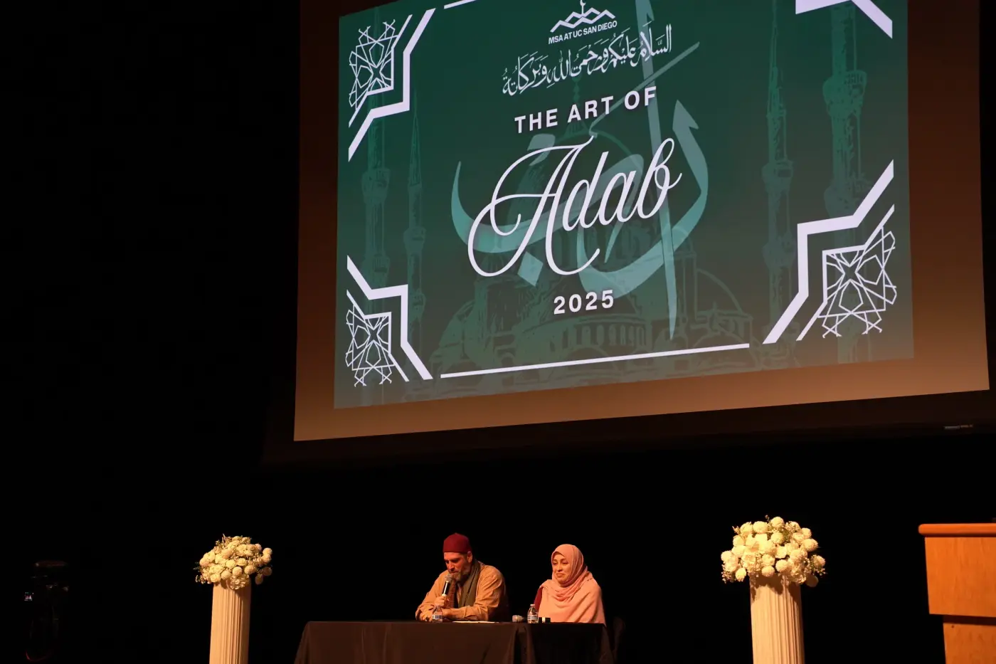 Two speakers seated at a table on stage for a panel beneath The Art of Adab slide