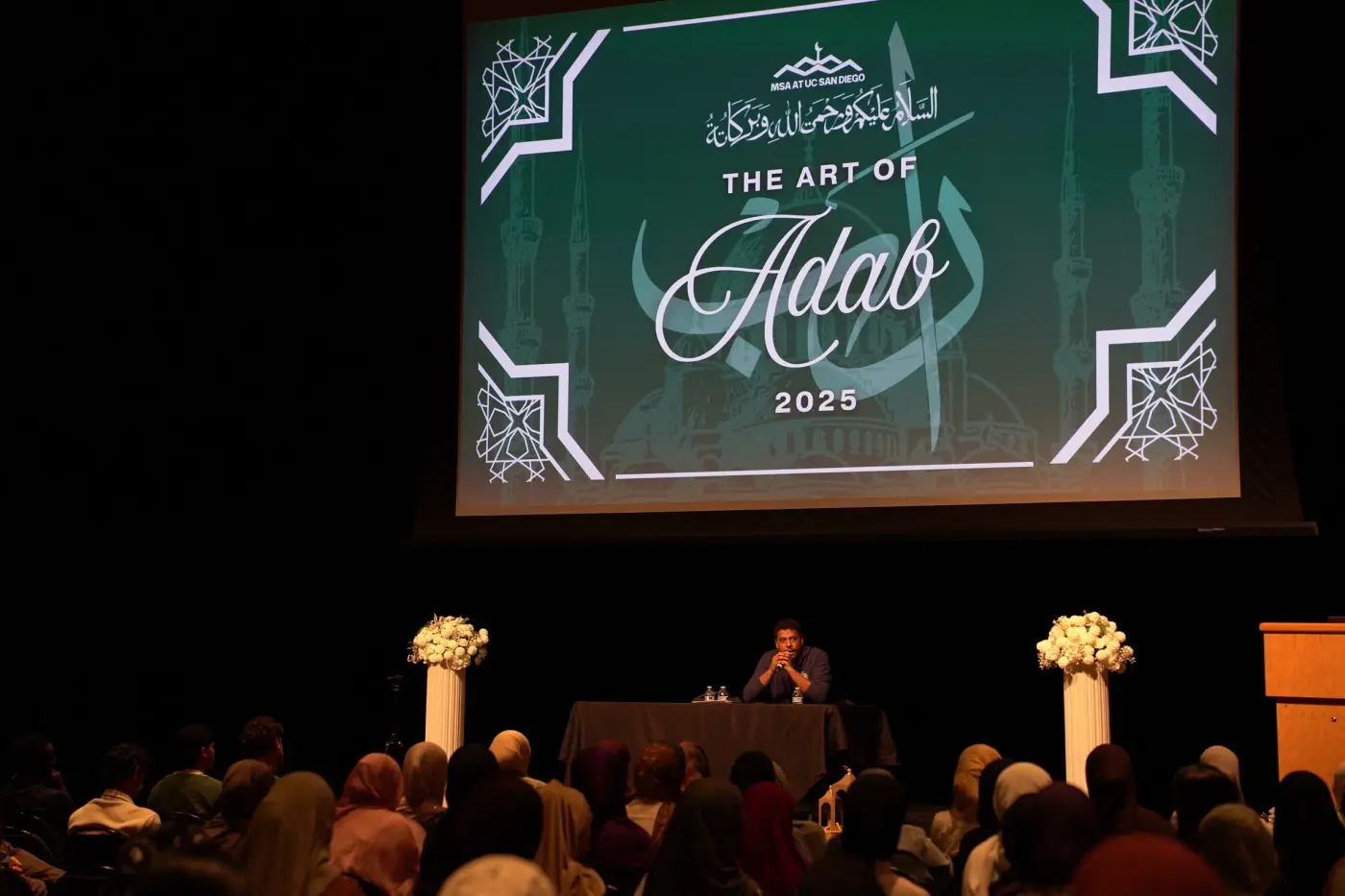 A full audience facing the stage beneath The Art of Adab 2025 slide while a speaker sits at the table