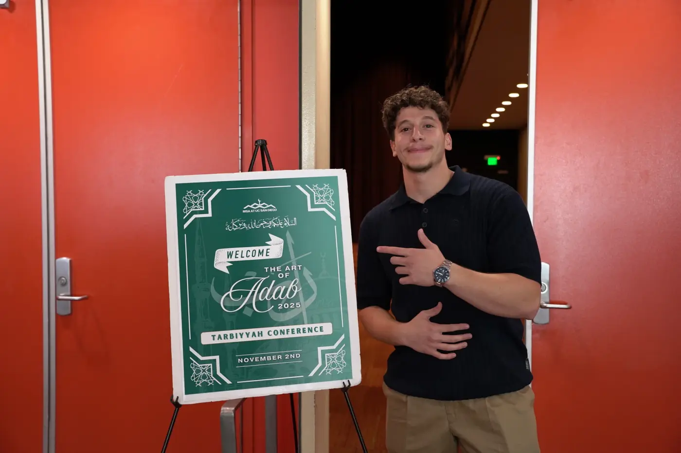 A volunteer gesturing to the green Art of Adab welcome sign at the hall entrance
